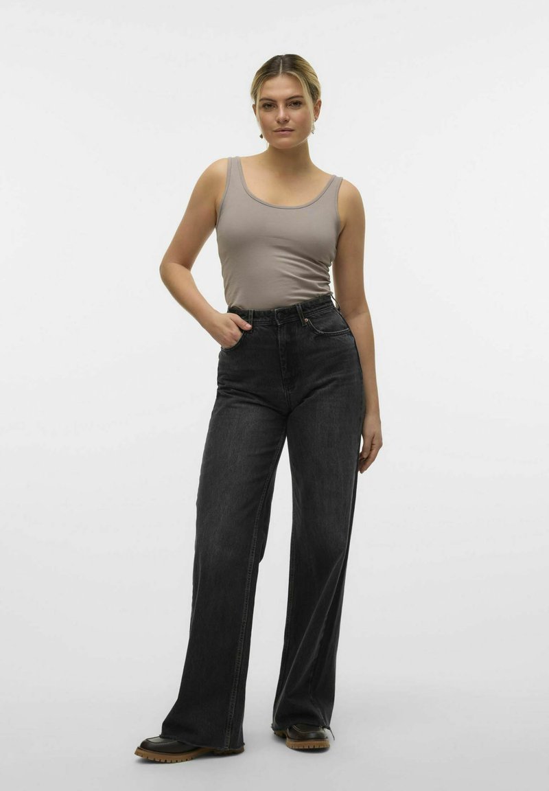 Szary tank top, czarne spodnie z wysokim stanem, szerokie nogawki, widoczne szwy i masywne czarne buty z brązowymi podeszwami. Jednolity biały tle.