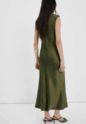 Femme aux longs cheveux noirs portant une robe midi sans manches de couleur vert olive et des talons noirs à lanières, debout devant un fond blanc.