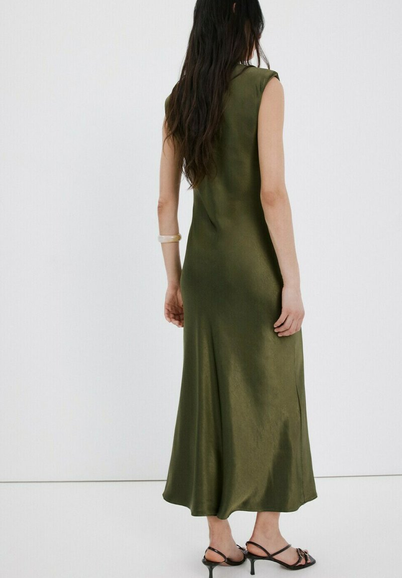 Žena s dlouhými tmavými vlasy má na sobě zelené midi šaty bez rukávů a černé sandály na podpatku, stojí před bílým pozadím.