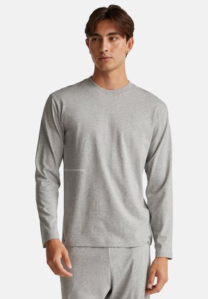 Joven vestido con una camiseta de manga larga gris lisa y pantalones grises a juego, de pie contra un fondo blanco.