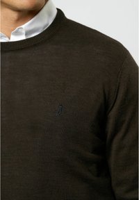 Temno zelena pletena jopica s okroglim izrezom, ki ima subtilno vezeno logo na levem prsnem delu in teksturirano tkanino.