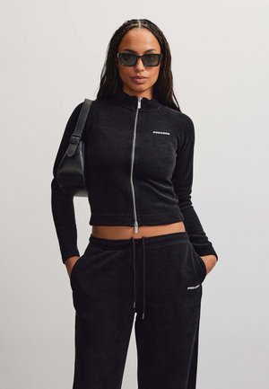 Femme portant une veste courte noire zippée et un pantalon assorti, les mains dans les poches, un sac à main noir à l'épaule et des lunettes de soleil foncées.