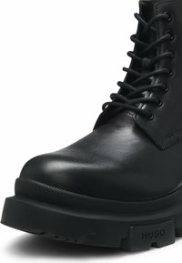 Black leather ankle boot with round toe, lace-up design, textured sole, and a prominent heel. Features a logo accent on the sole.