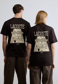 Μαύρο T-shirt με μεγάλο ταμπά γραφικό στην πλάτη, που περιλαμβάνει κείμενο και λογότυπα με θέμα το τένις. Κοντά μανίκια, σχέδιο με στρογγυλή λαιμόκοψη.