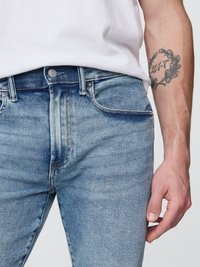 Jeans de denim azul desgastado con un corte ajustado, que cuentan con una cremallera, cinco bolsillos y un botón metálico visible. Textura ligera con variaciones sutiles.