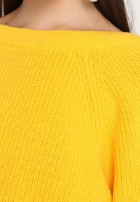 Jasnożółty sweter na drutach z szerokim dekoltem, z fakturowanym splotem i prążkowanymi wykończeniami przy mankietach i na dole.