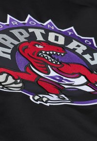 Tessuto nero con il logo dei Toronto Raptors. Il design include un dinosauro rosso con accenti bianchi e elementi di sfondo circolari viola.