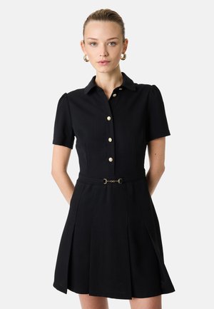 Kobieta w czarnej sukience z krótkim rękawem, z złotymi guzikami z przodu i wąskim paskiem, stojąca na jasnym, jednolitym tle.