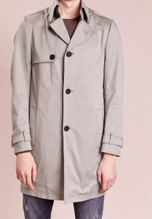 Mann trägt einen hellgrauen Trenchcoat mit schwarzen Knöpfen und verstellbaren Manschettenriemen über einem braunen Hemd und zerfetzten Jeans.