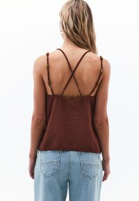 Top in tessuto marrone con una vestibilità ampia, dotato di spalline incrociate decorate con perline di colore ambra. Abbinato a jeans azzurri a vita alta.