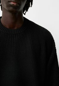 Pull noir tricoté avec une texture côtelée, présentant un col rond et une coupe ample. Le matériau semble doux et épais, conçu pour la chaleur.