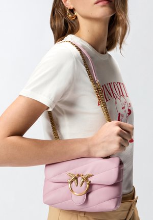 Femeie purtând un tricou alb cu grafică și pantaloni bej, având o geantă de umăr roz matlasată, cu încuietoare aurie în formă de pasăre și curea de lanț.