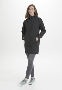 Musta vedenpitävä takki, jossa on vetoketjukiinnitys, säädettävä huppu ja sivutaskut. Päällä harmaat farkut ja harmaat urheilukengät.