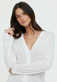 Cardigan en tricot blanc avec un motif en vagues, encolure en V et fermeture avec boutons. Texture douce avec des poignets et un ourlet côtelés.