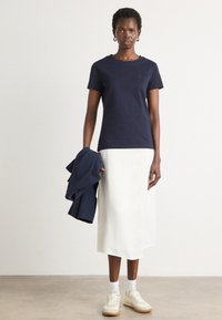 T-shirt in cotone blu marino con maniche corte, abbinato a una lunga gonna bianca. Scarpe casual e una giacca blu marino abbinata sono tenute nella mano sinistra.