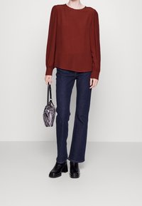Bordeauxfarbene Bluse mit langen Ärmeln und gerafften Schultern, kombiniert mit dunkelblauen Schlagjeans. Das Modell hält eine schwarz-weiße gemusterte Tasche. Schwarze Stiefel mit Absatz.