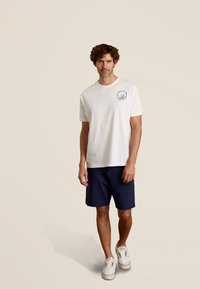 Witte katoenen T-shirt met een klein multikleurig zeilbootlogo, gecombineerd met marineblauwe shorts en witte sneakers. Model staat op een neutrale achtergrond.