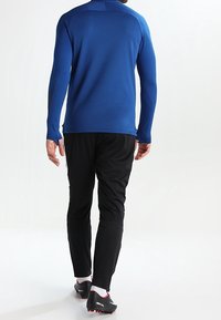 Maglia atletica a maniche lunghe di colore blu reale abbinata a pantaloni neri. Presenta un design aderente, una texture liscia e calzature nere con dettagli rossi.