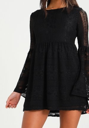 Femme portant une robe mini en dentelle noire avec de longues manches évasées et un col montant, se tenant devant un fond clair.