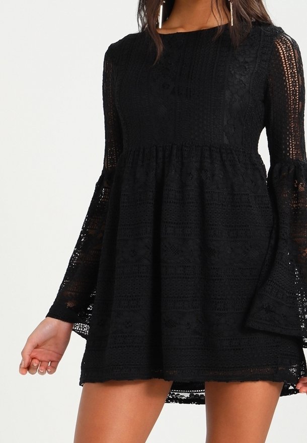 Kvinna iklädd en svart spets minikjol med långa trumpetärmar och hög krage, stående mot en enfärgad ljus bakgrund.