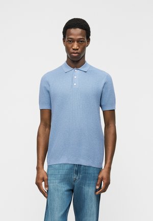 Tânăr în picioare, purtând un tricou polo cu mâneci scurte de culoare albastru deschis și blugi albastru mediu, pe un fundal simplu, deschis.