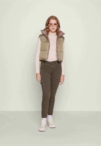 Puffer vest v olivově zelené barvě, pletený svetřík v jemně růžové barvě, olivové cargo kalhoty, bílé ponožky a tenisky. Model stojí proti světlému zelenému pozadí.