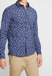 Camicetta a righe navy con motivi a foglia azzurri, colletto button-down, maniche lunghe e orlo curvo, abbinata a pantaloni beige.