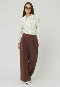 Blouse blanche à motifs floraux avec un col volanté, associée à un pantalon large taille haute marron et des baskets blanches. Lunettes de soleil incluses.
