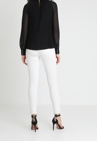 Blusa nera plissè con aperture a chiave sul retro, abbinata a jeans skinny bianchi e sandali neri con tacco alto dotati di dettagli sulla caviglia.