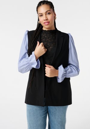 Zwarte mouwloze blazer met gestructureerde pasvorm, gecombineerd met een lichtblauwe gestreepte blouse met pofmouwen en een zwarte kanten top eronder.