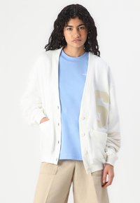 Cardigã branco com um ajuste solto, fecho de botão e dois bolsos frontais, sobre uma t-shirt azul clara com "NEW" estampado.