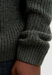 Dark green knitted sweater fabric with ribbed texture, showcasing a cuff and hand in a smooth brown tone. Close-up detail of material.