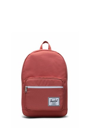 Sac à dos en tissu rouge avec une poche zippée à l'avant, des bretelles réglables et une poignée supérieure. Présente une étiquette blanche à l'avant avec le nom de la marque.