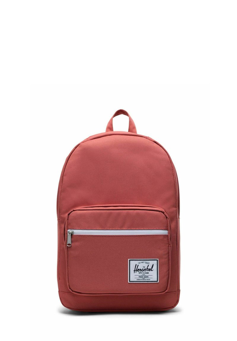 Sac à dos en tissu rouge avec une poche zippée à l'avant, des bretelles réglables et une poignée supérieure. Présente une étiquette blanche à l'avant avec le nom de la marque.