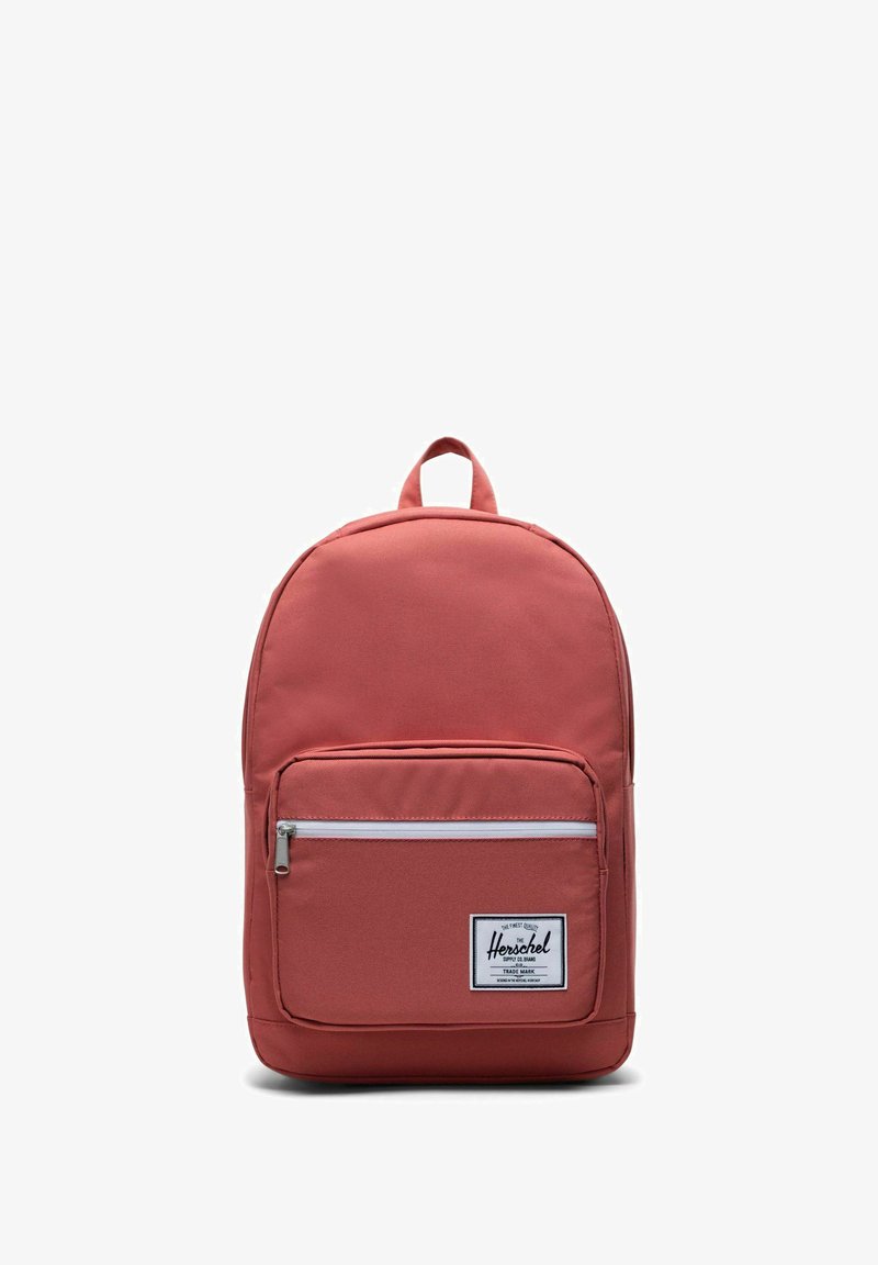 Sac à dos en tissu rouge avec une poche zippée à l'avant, des bretelles réglables et une poignée supérieure. Présente une étiquette blanche à l'avant avec le nom de la marque.