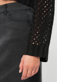 Pull en maille noire avec des manches ajourées, doté d'un poignet côtelé. Porté sur un jean gris foncé, mettant en valeur un tissu texturé et une coupe ajustée.