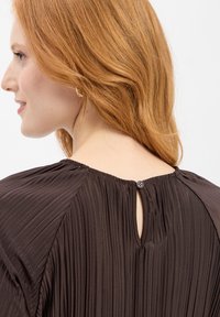 Brun plisserad blus med nyckelhålsstängning bak, rund halsringning och mjuk drapering. Mjuk textur och subtil glans.