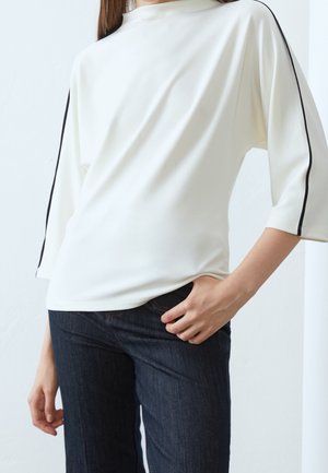 Femme portant un haut blanc à manches longues avec une bande noire sur les manches et un jean bleu foncé, main dans la poche sur fond blanc.