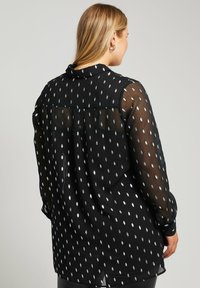 Zwarte doorschijnende blouse met een zilveren stippenpatroon, lange mouwen, een kraag en knoopmanchetten. De achterkant heeft een licht losse pasvorm.