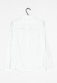 Bílá košile s dlouhým rukávem a modrým květinovým vzorem, s špičatým límcem a knoflíkovými manžetami, vystavená na černém ramínku.