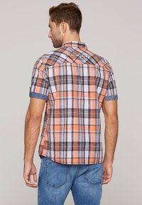 Lyhythihainen ruudullinen paita oranssina, mustana ja valkoisena, jossa on napitettavat kaulukset ja denimiä hihoissa. Merkin logo takana.