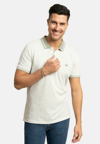 Polo de color gris claro con un patrón texturizado y acentos de color verde en el collar, combinado con jeans de mezclilla. La camisa presenta un pequeño logo en el pecho.