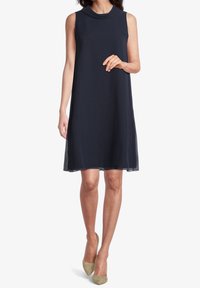 Sleeveless, knee-length navy dress with a high neck, soft fabric, and subtle sheer hem, paired with light green pointed-toe heels.