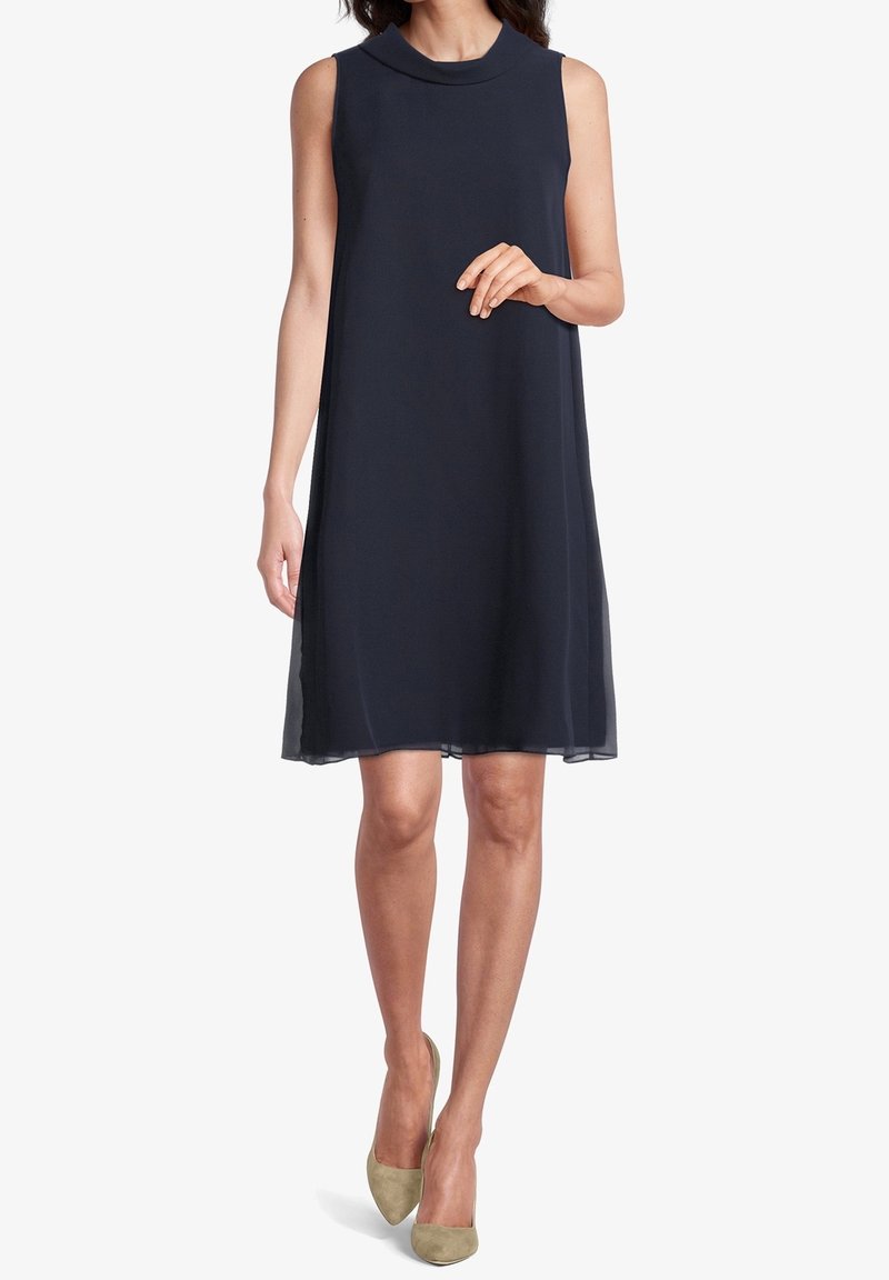 Sleeveless, knee-length navy dress with a high neck, soft fabric, and subtle sheer hem, paired with light green pointed-toe heels.