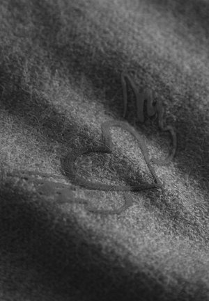 Grijze stof met een zachte, fuzzy textuur, featuring een gestikt logo in donker, reliëfleer. Het ontwerp omvat een hartvorm en gestileerde letters.