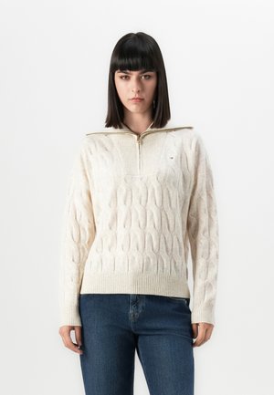 Woman with straight dark hair wears a cream cable-knit sweater with a front zipper and blue jeans against a plain white background.
