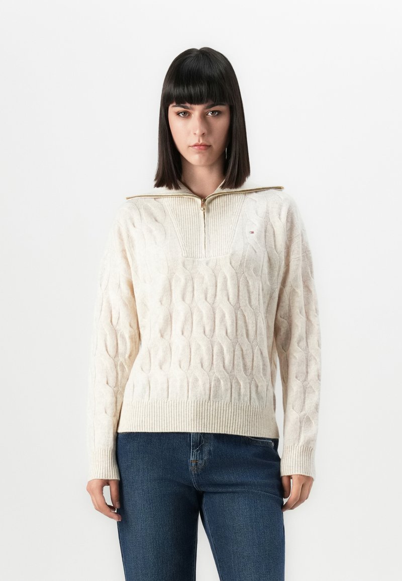 Une femme avec des cheveux raides et foncés porte un pull en tricot torsadé crème avec une fermeture éclair à l'avant et un jean bleu sur un fond blanc uni.