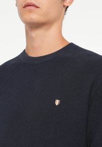Marineblå teksturert genser med rund hals, som har et lite skjoldlogo med vertikale striper i krem og brunt på venstre bryst.
