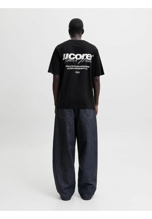 Personne debout de dos, portant un T-shirt noir avec un texte blanc et un jean large bleu foncé sur un fond clair uni.
