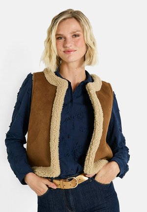 Femme blonde avec des cheveux ondulés portant un gilet en shearling brun par-dessus un haut brodé bleu marine, un jean bleu et une ceinture beige, les mains dans les poches.